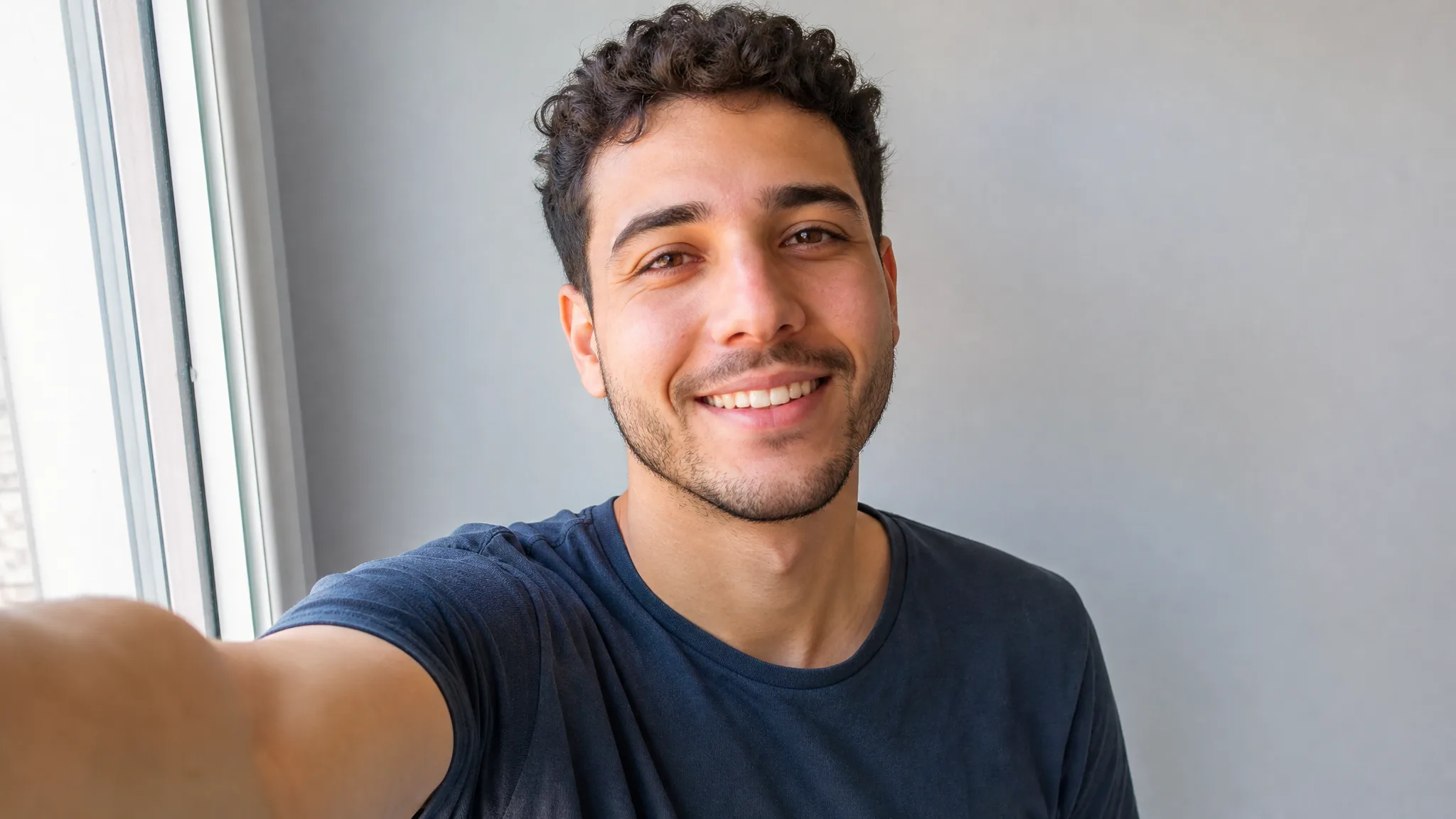 The Selfie That Starts It A curly-haired young man in a navy tee smiles at his outstretched phone in daylight by a window