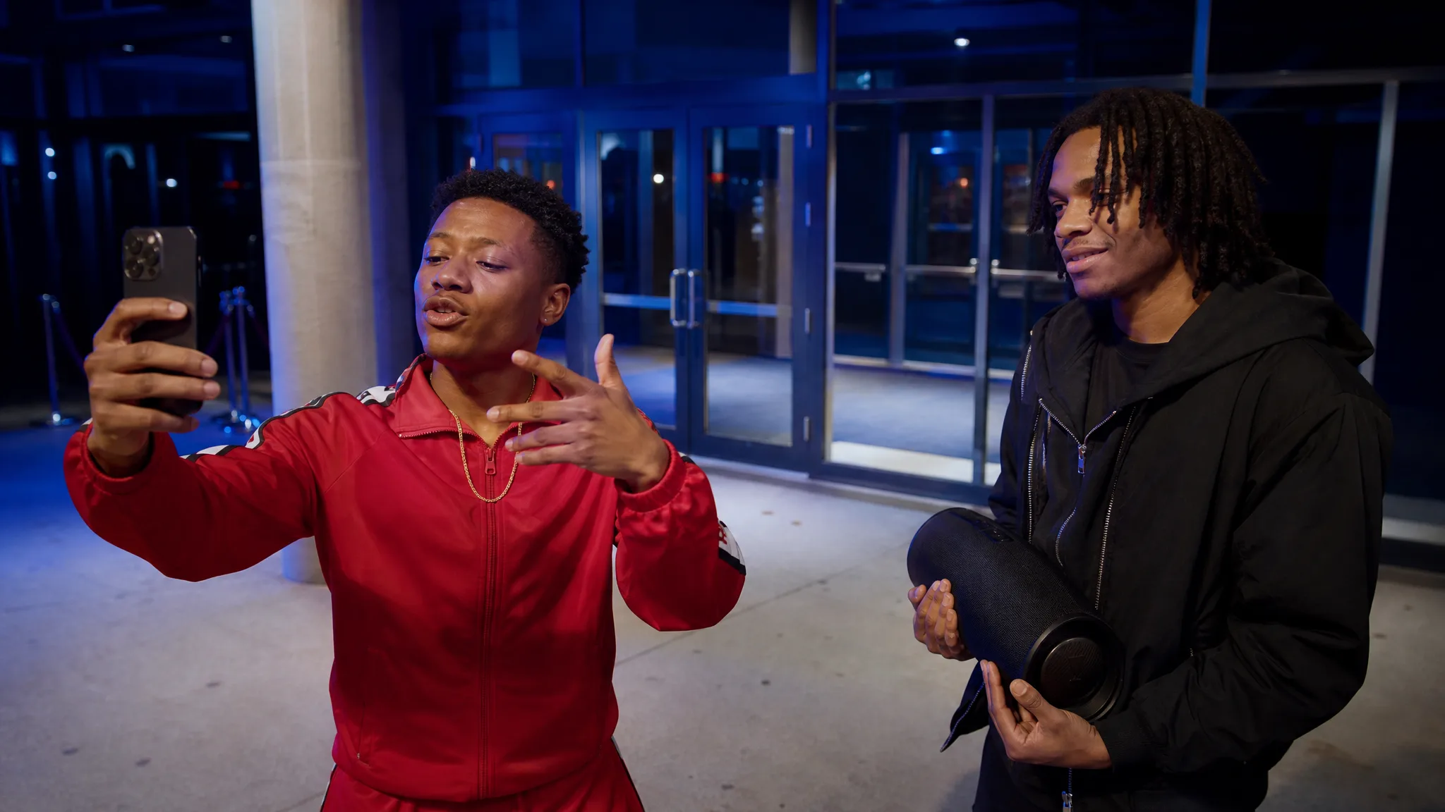 Red Tracksuit and Gold Chain A young man in a red tracksuit and thin gold chain raps at his phone outside a blue-lit glass door, a friend holding a speaker